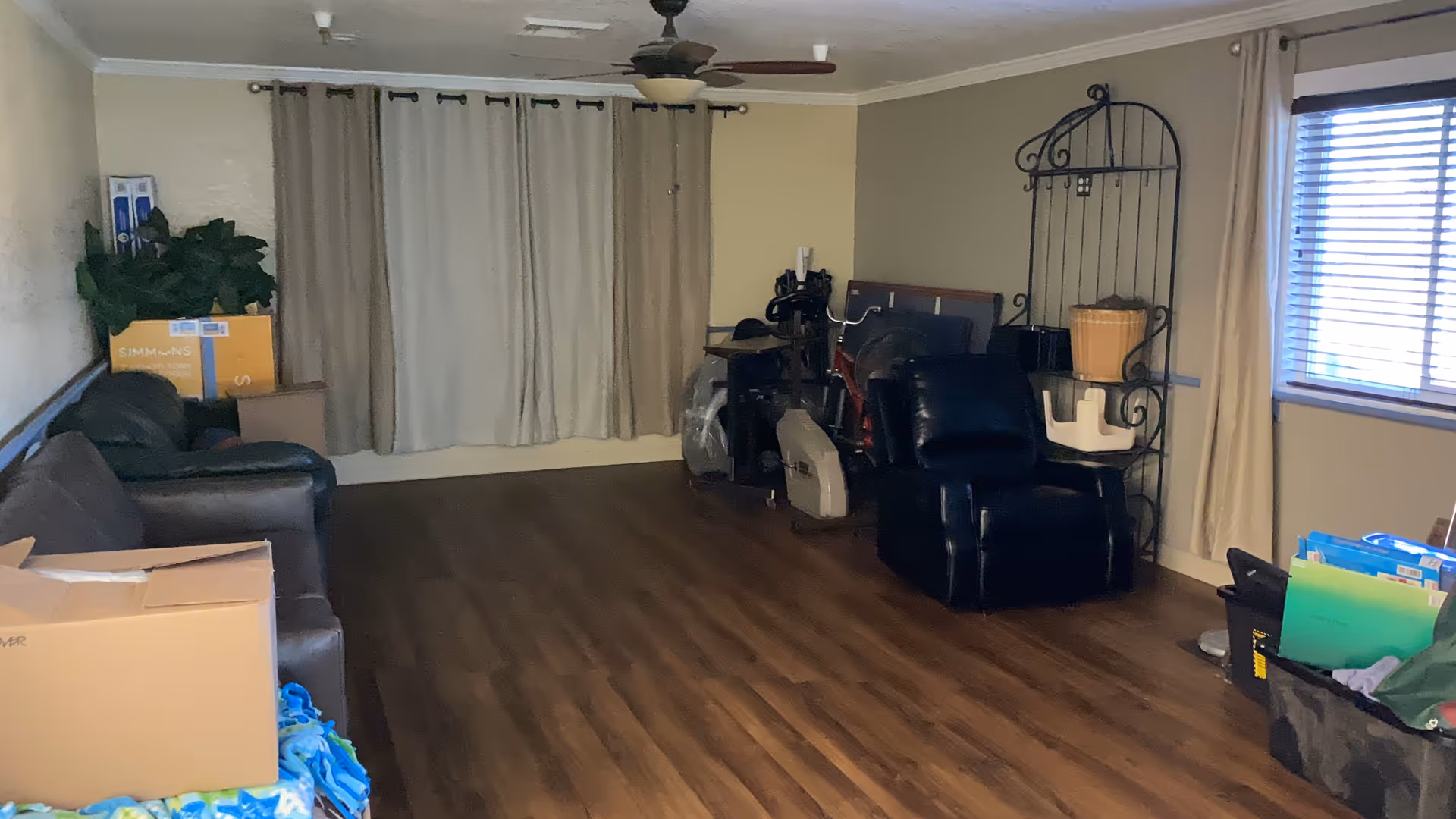 A dimly lit living room with wood flooring, a ceiling fan, and beige walls. The room contains a black leather recliner, a green couch, a metal shelf with a basket, some boxes, and various items scattered around, including a bicycle and exercise equipment near the back wall with closed curtains.