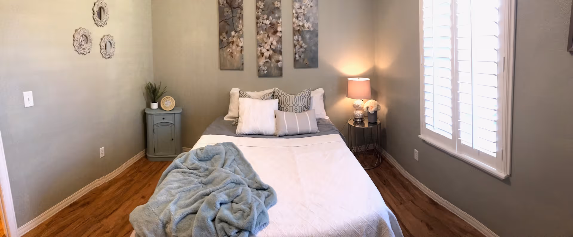 A cozy bedroom with a bed centered against the back wall, adorned with multiple pillows and a light blue throw blanket. On the left side of the bed is a small green nightstand with a plant and decorative plate, and on the right side is a round table with a lamp and flower arrangement. The walls are painted a soft green, decorated with three floral paintings above the bed and three small decorative wall hangings on the left wall. A window with white plantation shutters is on the right wall, and the floor is wooden.
