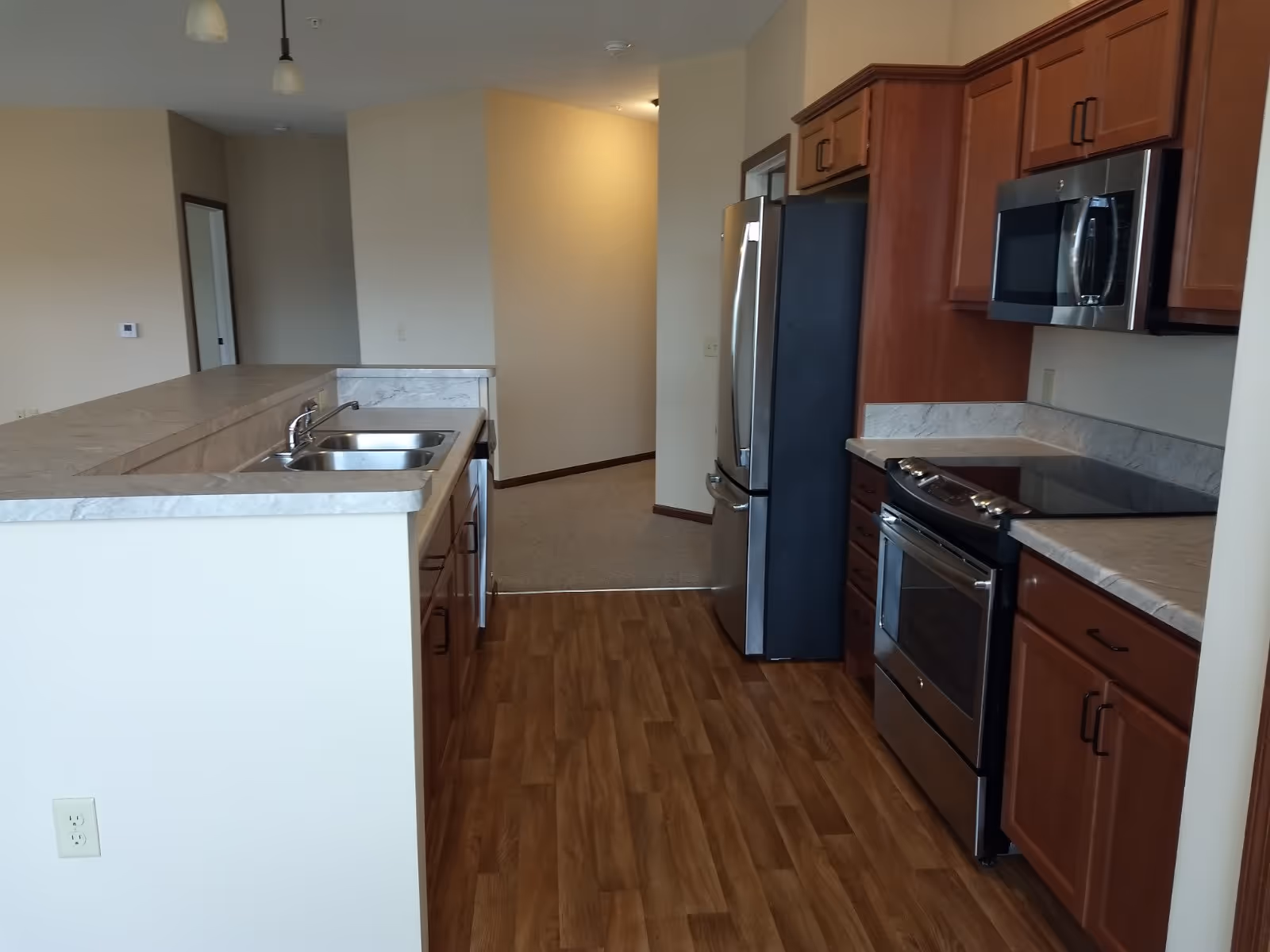 Open modern kitchen with stainless steel refrigerator, stove and microwave, a double sink in a breakfast bar, wooden cabinets and wood-look flooring.