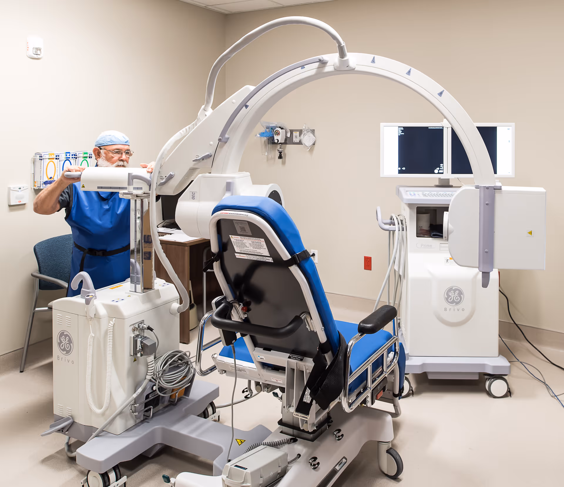 A medical professional wearing blue scrubs and a surgical cap is adjusting a large piece of medical imaging equipment in a clinical room. The equipment includes a blue and black patient chair and a GE Brivo imaging machine with monitors displaying medical images. The room has beige walls and medical supplies on the wall.