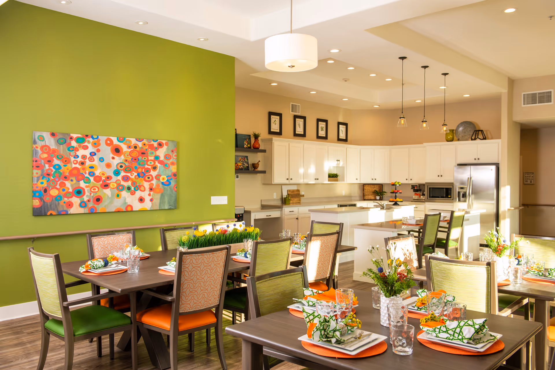 A bright and modern dining area with several tables set for a meal, featuring colorful napkins and floral centerpieces. The room has a green accent wall with a vibrant abstract painting. In the background, there is a spacious kitchen with white cabinets, stainless steel appliances, and pendant lighting.