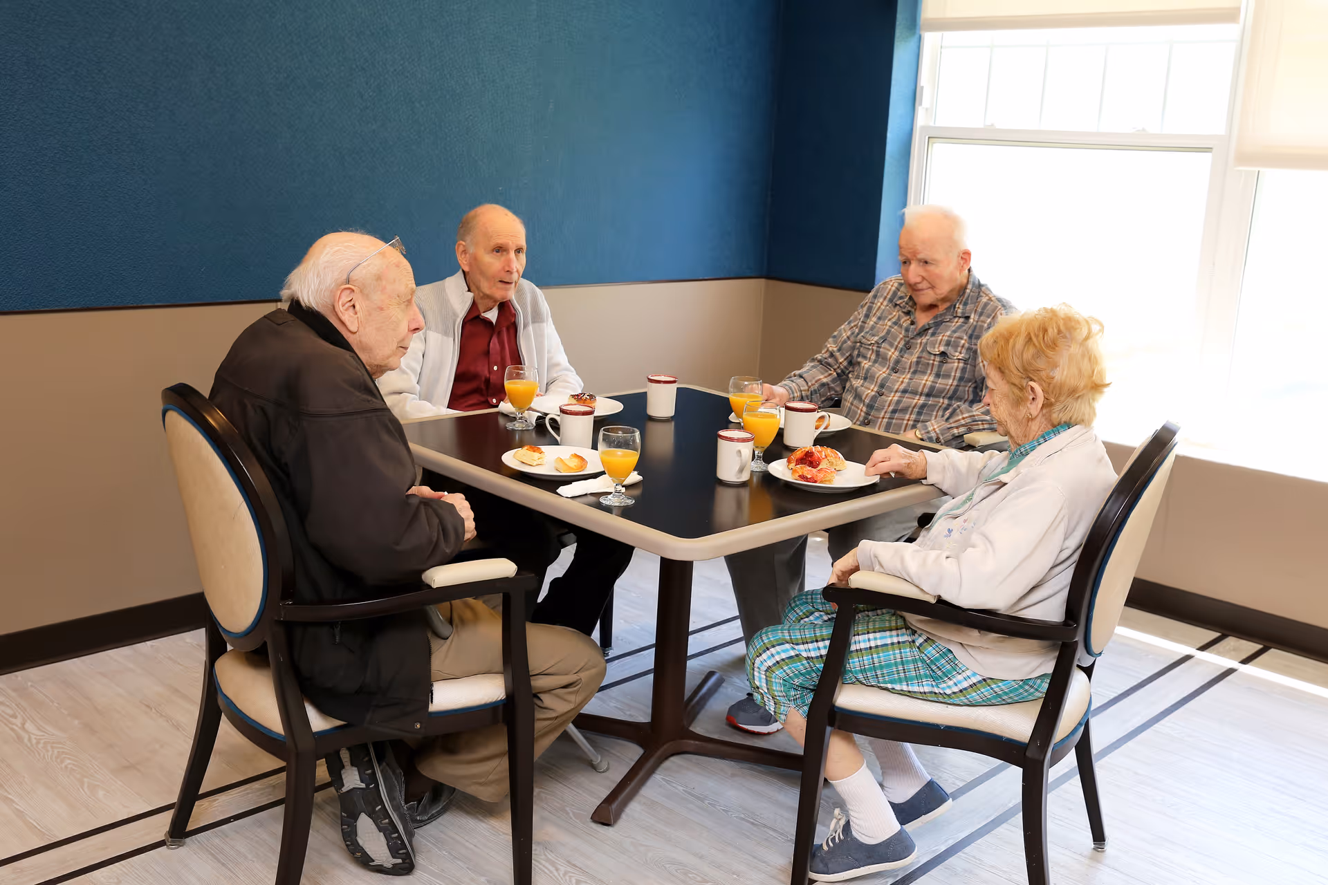 Four elderly people sitting around a square dining table in a well-lit room with blue and beige walls. They are having a meal with plates of food, glasses of orange juice, and coffee mugs on the table.