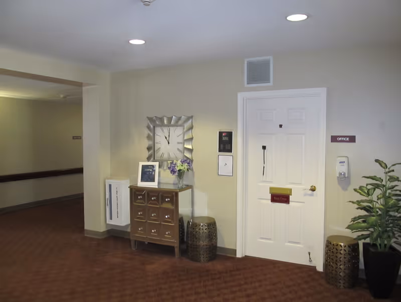 Interior hallway of a senior living facility with a white door labeled 'Office'. Next to the door is a hand sanitizer dispenser mounted on the wall and a potted plant. A small chest of drawers with a clock, framed picture, and a vase of flowers sits against the wall. Two decorative stools are placed near the door and chest. The floor is carpeted and the walls are painted light beige.