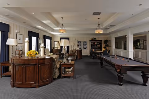 A spacious and elegant common area with a pool table on the right, multiple seating arrangements with sofas and armchairs, wooden tables with lamps and decorative items, and large windows with dark curtains. The room features a coffered ceiling with hanging light fixtures and a carpeted floor.