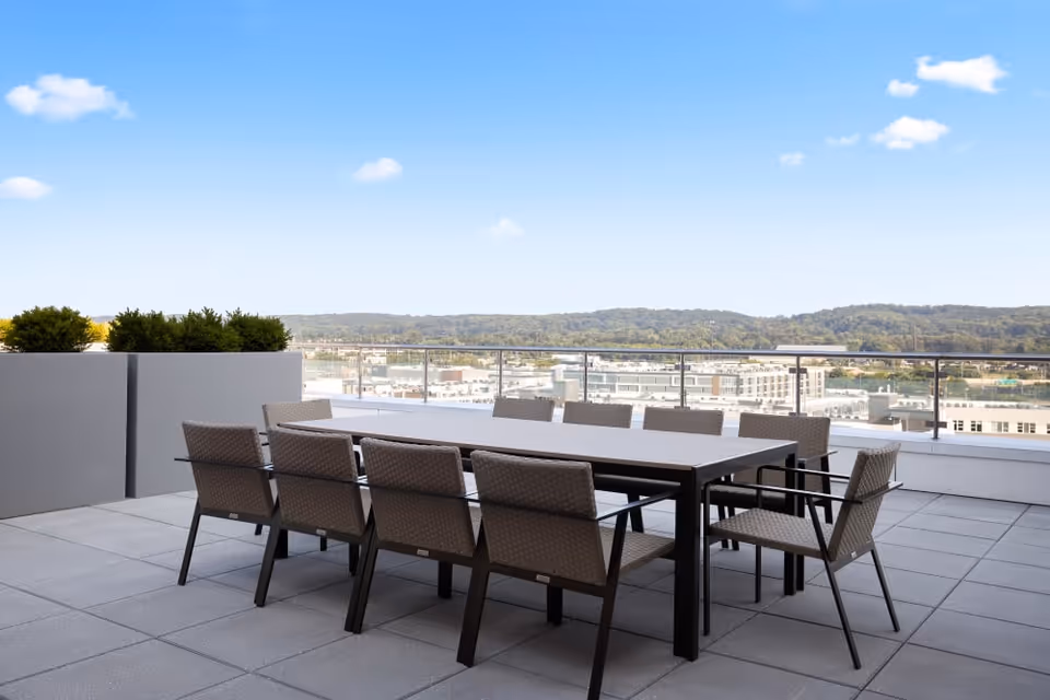 Outdoor patio area with a long rectangular table surrounded by ten woven chairs. The patio has large square tiles and glass railing, overlooking a cityscape with hills in the background under a blue sky with a few clouds.