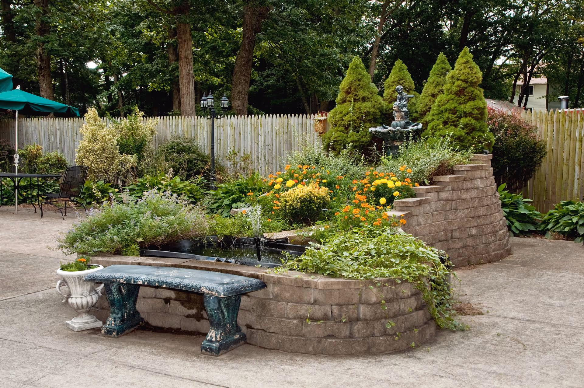 A peaceful outdoor garden area with a curved stone planter filled with various green plants and colorful flowers. There is a small water fountain with a statue in the center of the planter. A green stone bench and a white planter with flowers are placed in front of the planter. In the background, there is a wooden fence, tall trees, and a black metal table with chairs under green umbrellas.