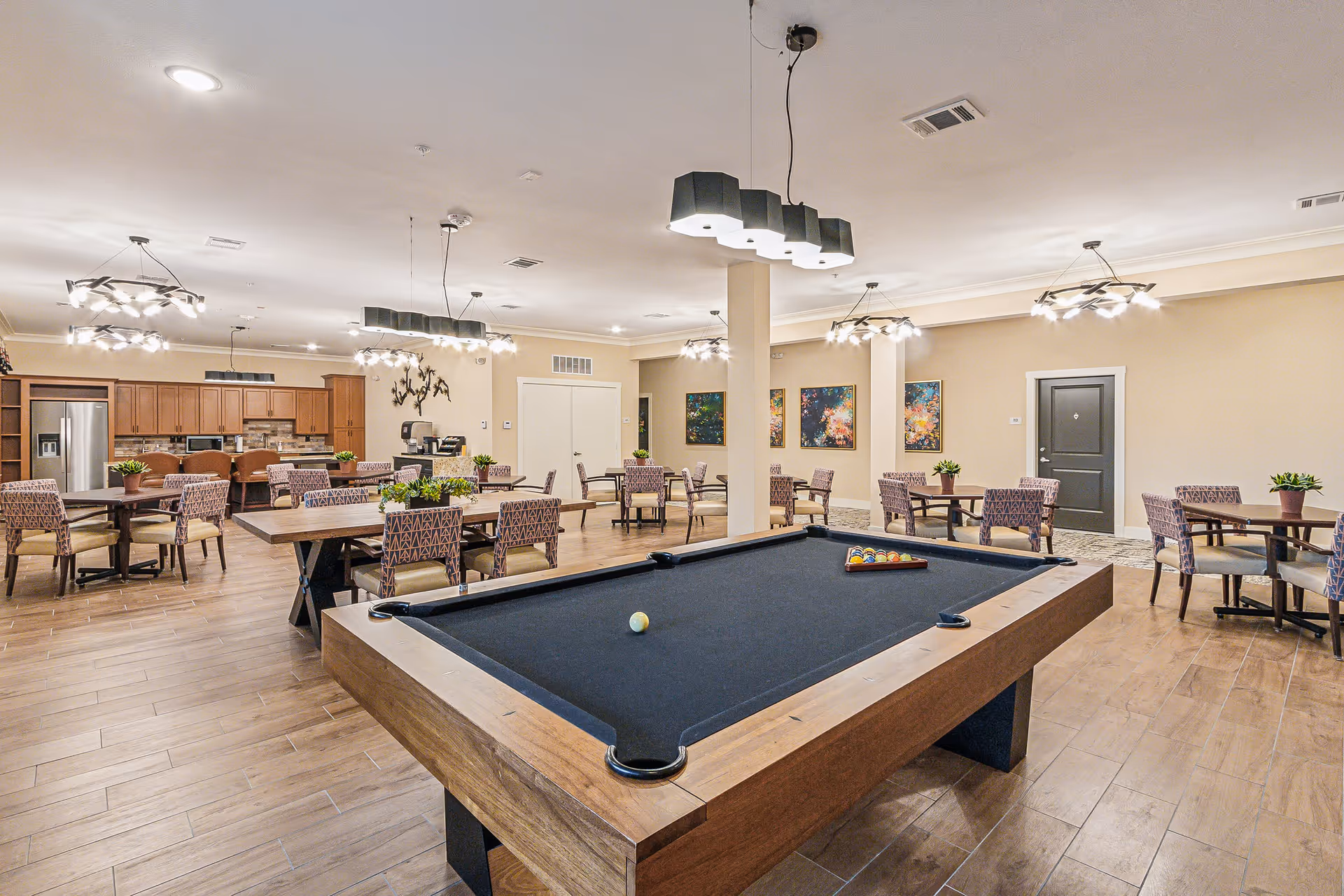 A spacious senior living common room with a pool table in the foreground and dining tables with a kitchenette in the background.