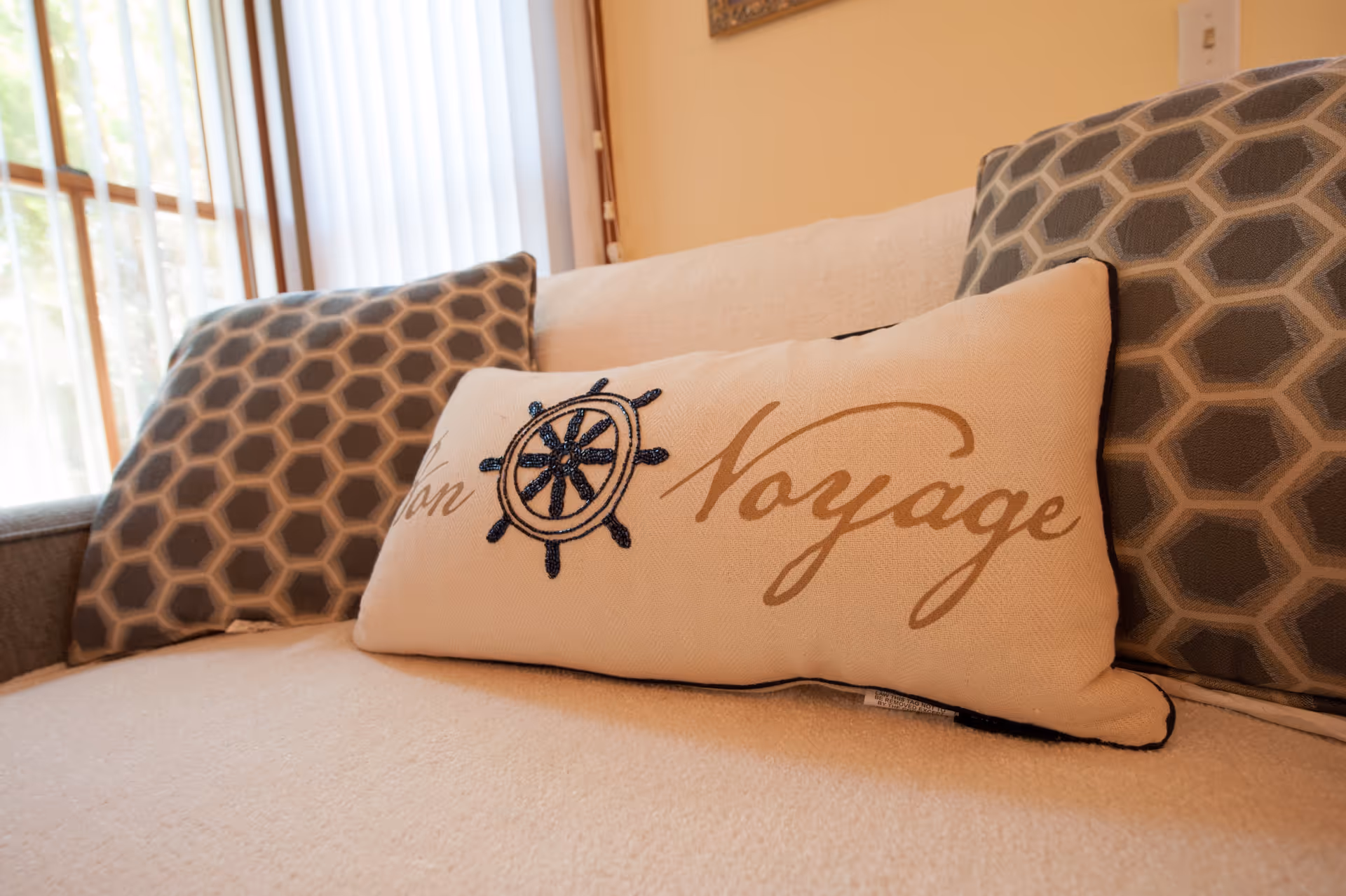 Close-up of a beige couch with two gray hexagon-patterned pillows and a decorative rectangular pillow in the center that has a ship's wheel design and the words 'Bon Voyage' embroidered on it. A window with vertical blinds is visible in the background.
