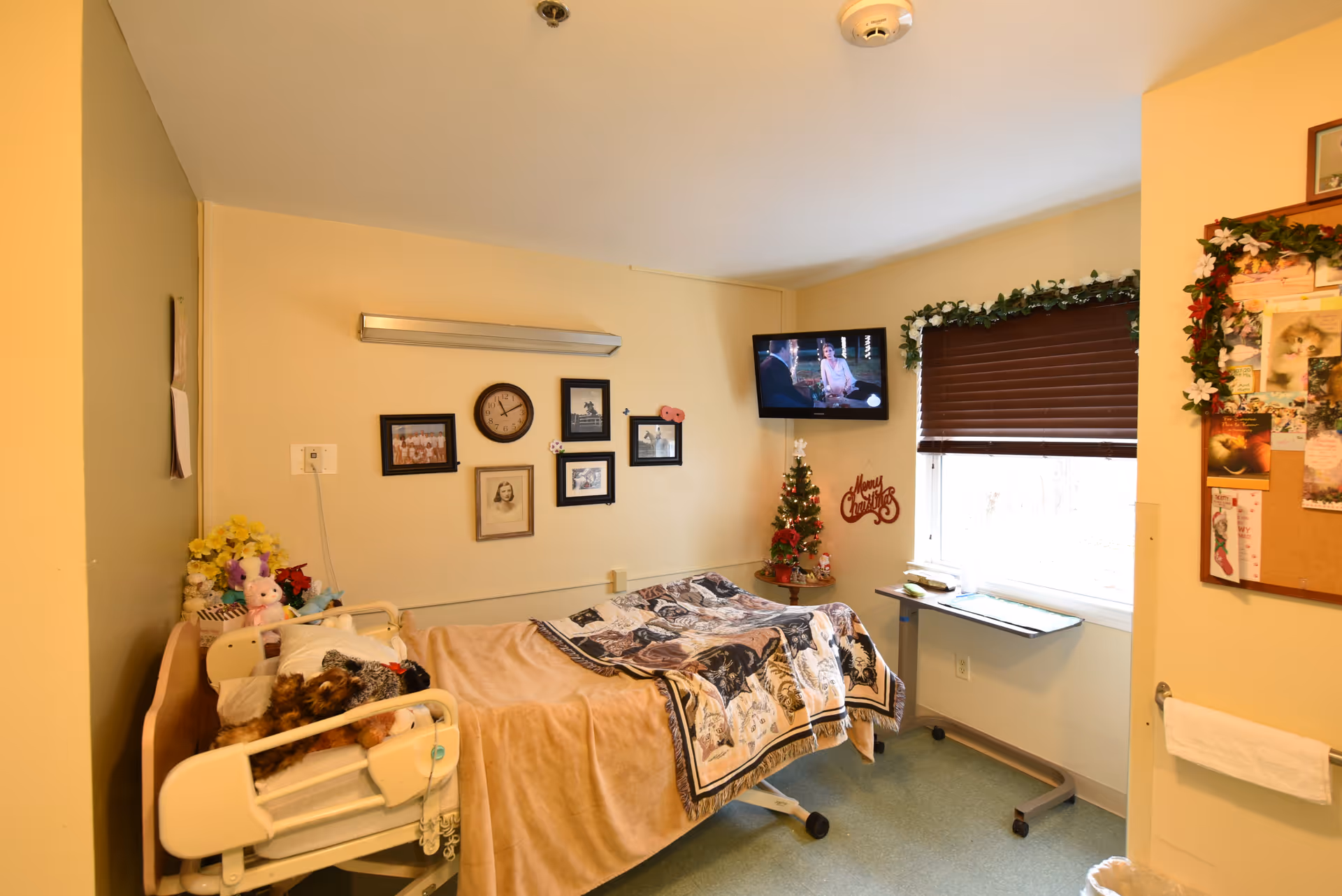 A cozy nursing facility room with a single bed covered in a beige blanket and a patterned quilt. The bed has stuffed animals on it. On the wall above the bed are several framed photos and a clock. A small decorated Christmas tree sits on a table near a window with brown blinds and garland. A flat-screen TV is mounted on the wall showing a scene from a movie or show. A bulletin board with various notes and pictures is on the right wall, and a towel hangs below it.