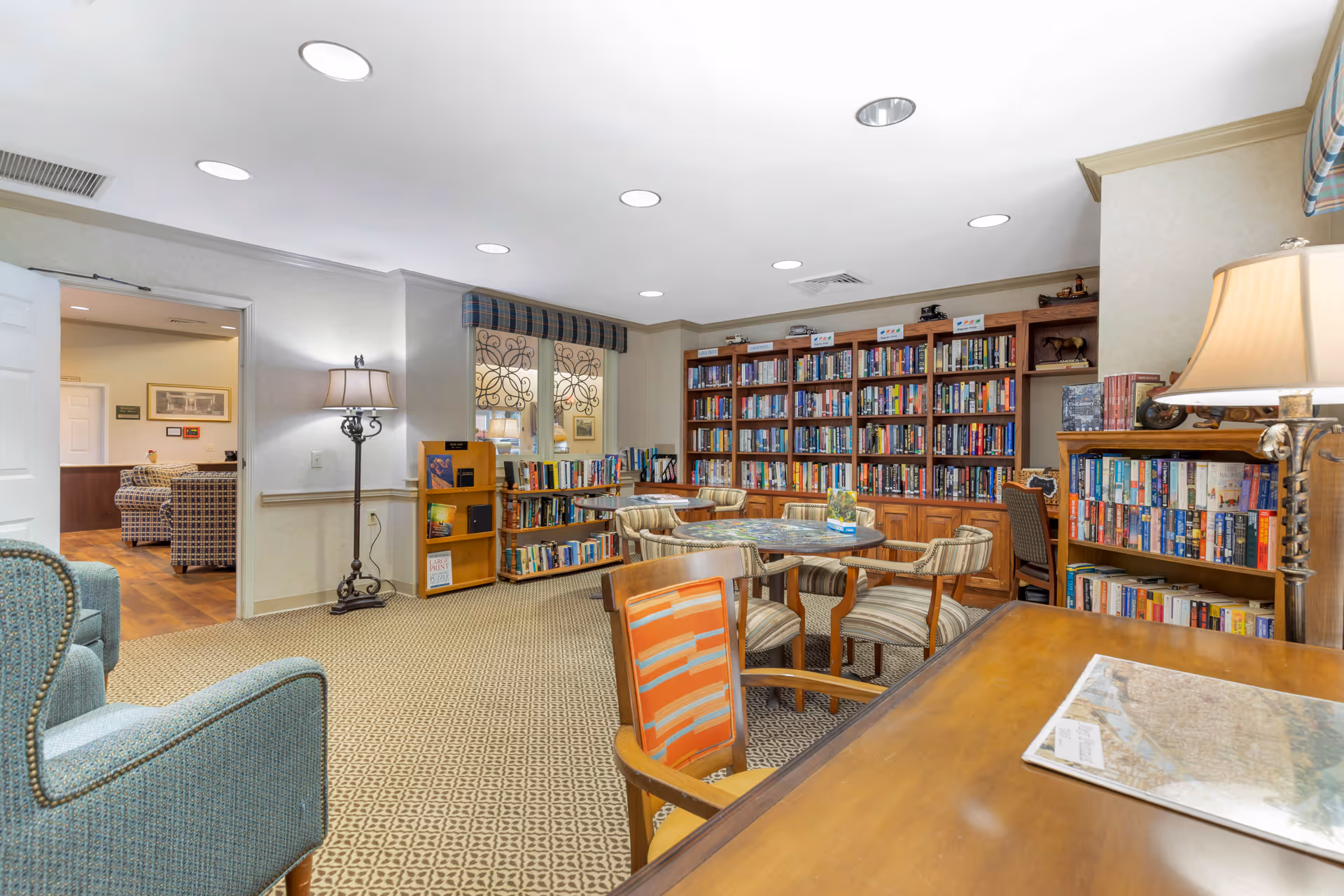 A cozy senior living facility library room with bookshelves filled with books along the walls, a round table surrounded by striped cushioned chairs, a wooden desk with a lamp, and a comfortable armchair. The room is well-lit with recessed ceiling lights and has a patterned carpet. An open door reveals another sitting area with plaid upholstered chairs.