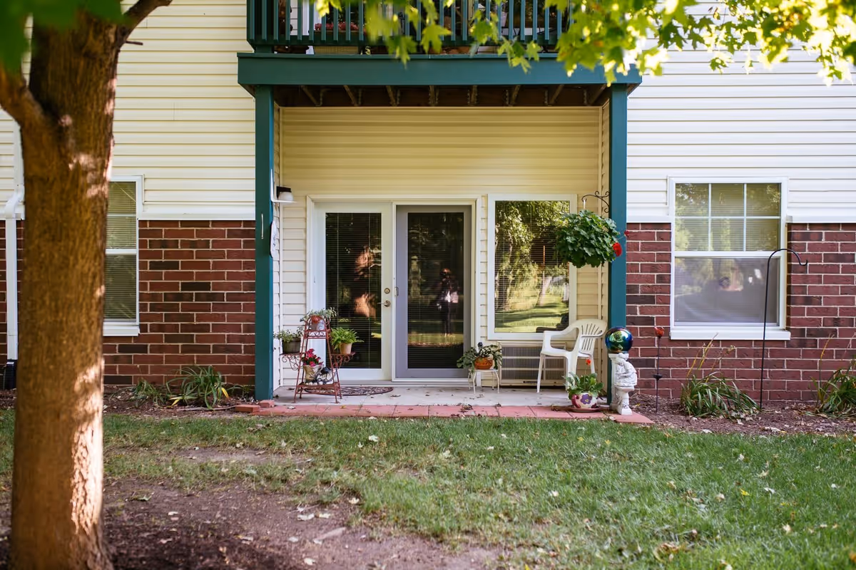 Outdoor patio area of a residential building with a glass door and window. The patio has potted plants, a white plastic chair, and a small decorative statue. The building exterior features a combination of brick and light-colored siding. A tree and green grass are visible in the foreground.