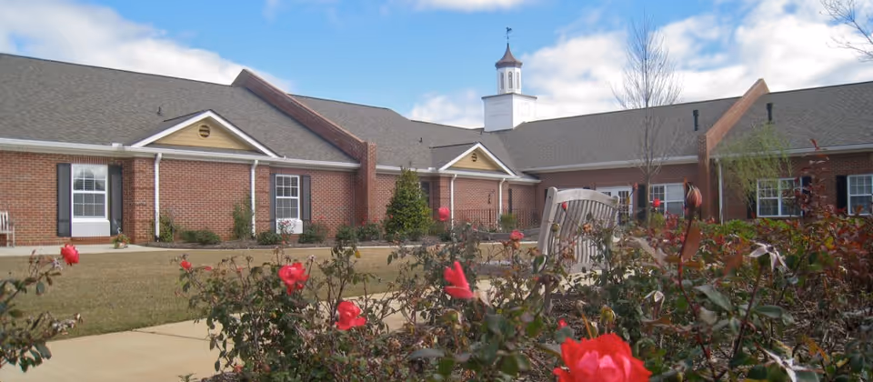 Outdoor view of Ashlan Village showing a single-story brick building with white-trimmed windows and a cupola on the roof. In the foreground, there is a garden with blooming red roses and a wooden bench along a paved walkway. The sky is partly cloudy.