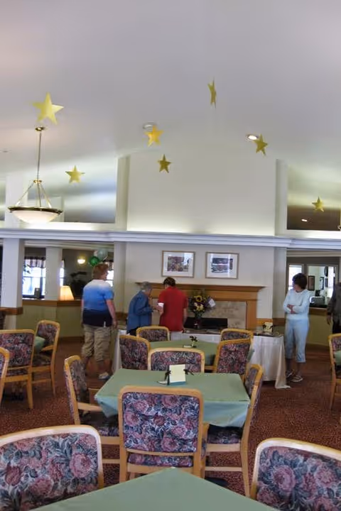 Communal dining room with floral-upholstered chairs and tables, a fireplace, hanging star decorations, and several people standing near the back.