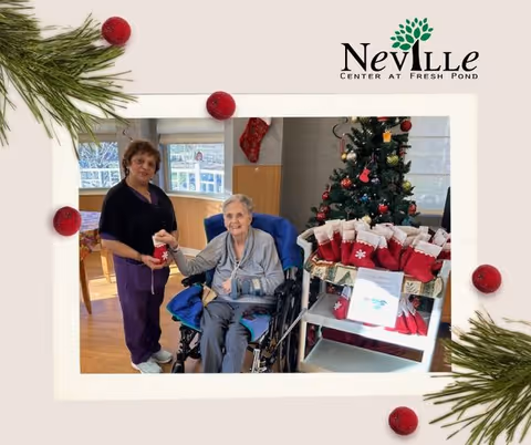An elderly woman sitting in a wheelchair holding hands with a caregiver standing beside her in a festive room decorated for Christmas. There is a Christmas tree adorned with ornaments and a display of red and white Christmas stockings on a cart nearby. The room has large windows and wooden flooring.