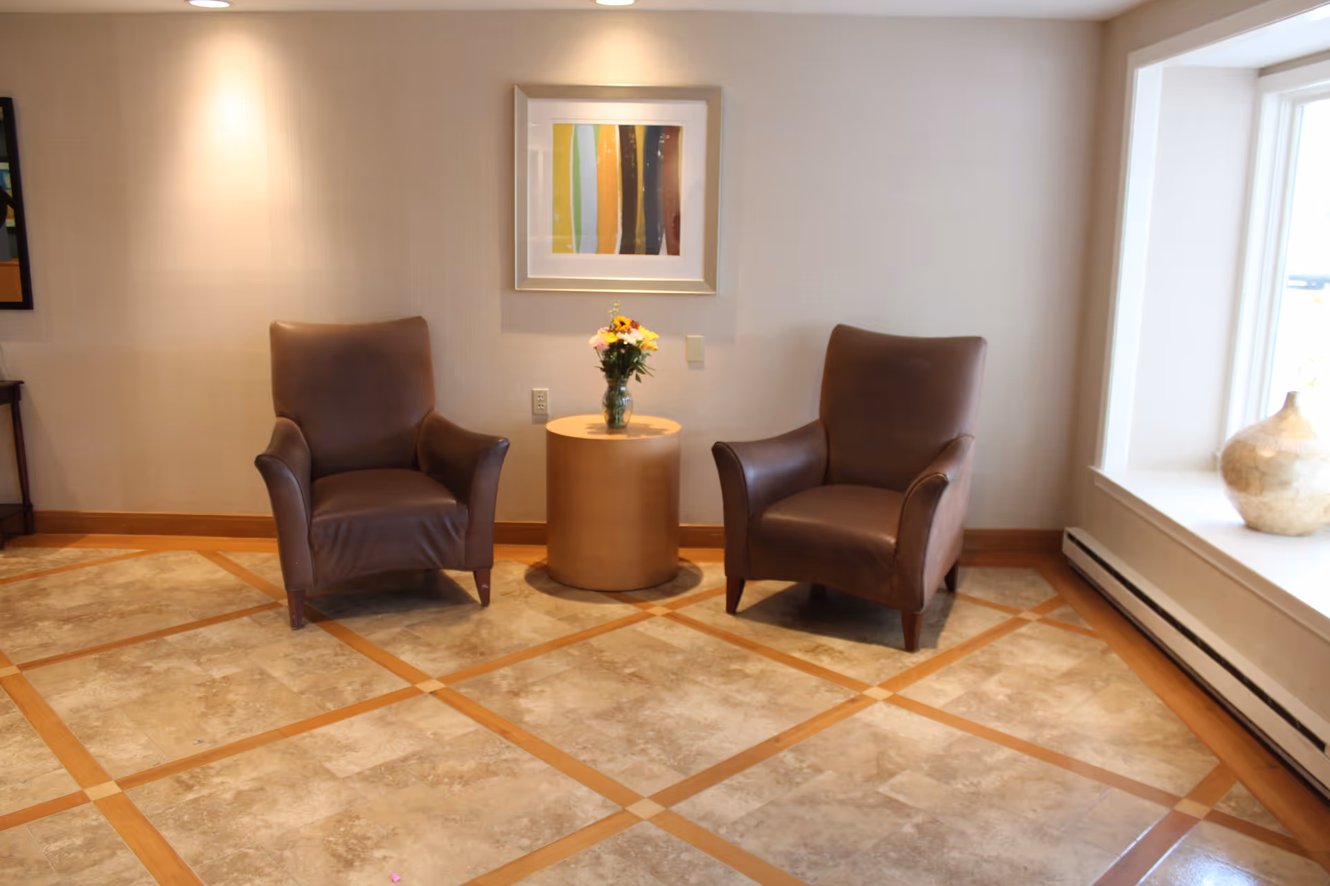 Two brown leather armchairs with a small round wooden table between them holding a vase of flowers, set against a beige wall with a framed abstract painting above. The floor has a tile pattern with wooden inlays, and there is a large window with a decorative vase on the windowsill to the right.