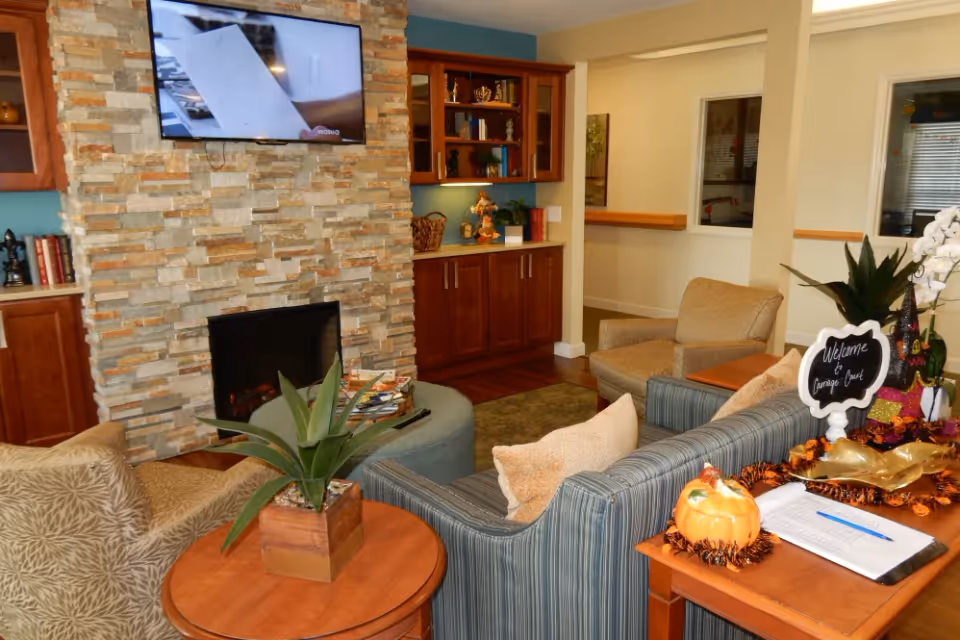 Cozy common living room with sofas and armchairs arranged around a stone fireplace and wall-mounted TV, with a side table displaying a 'Welcome to Carriage Court' sign and seasonal decorations.