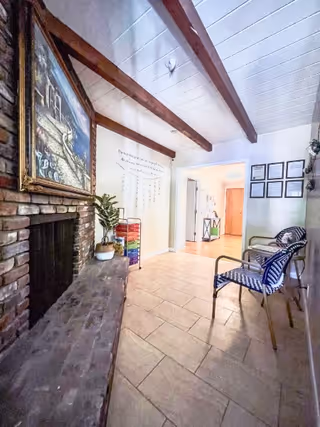 Cozy interior common area with a brick fireplace and bench, exposed wooden ceiling beams, chairs along a wall, and decorative artwork.