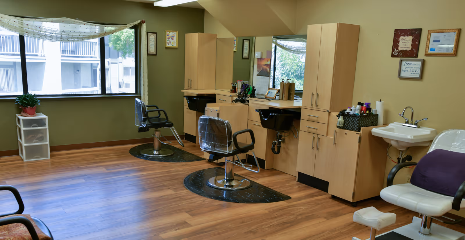 Interior of a salon area in a senior living facility with two salon chairs in front of mirrors and washbasins, a white chair with a purple cushion, wooden flooring, a large window with sheer curtains, and framed inspirational quotes on the wall.
