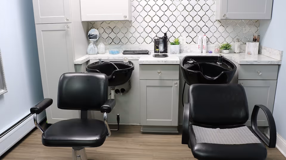 Two black salon shampoo chairs and basins in front of gray cabinets and a patterned tile backsplash.