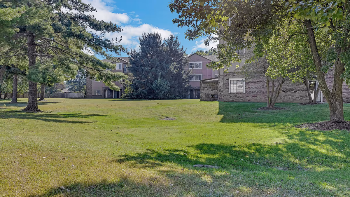 A wide grassy lawn with scattered trees in front of a multi-story brick senior living building under a partly cloudy sky.