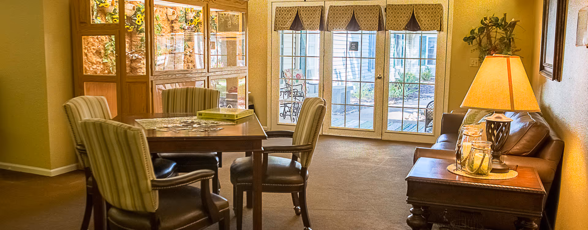 Cozy common room with a dining table and chairs, a leather sofa and side table with a lamp, and glass doors opening to a patio.