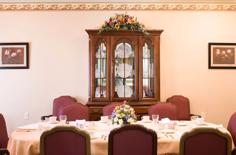 A dining room with a table set for a meal, featuring a floral centerpiece, cups, glasses, and utensils. The table is surrounded by maroon chairs, and a wooden china cabinet with plates and glassware is against the wall. Two framed floral paintings hang on the beige walls.