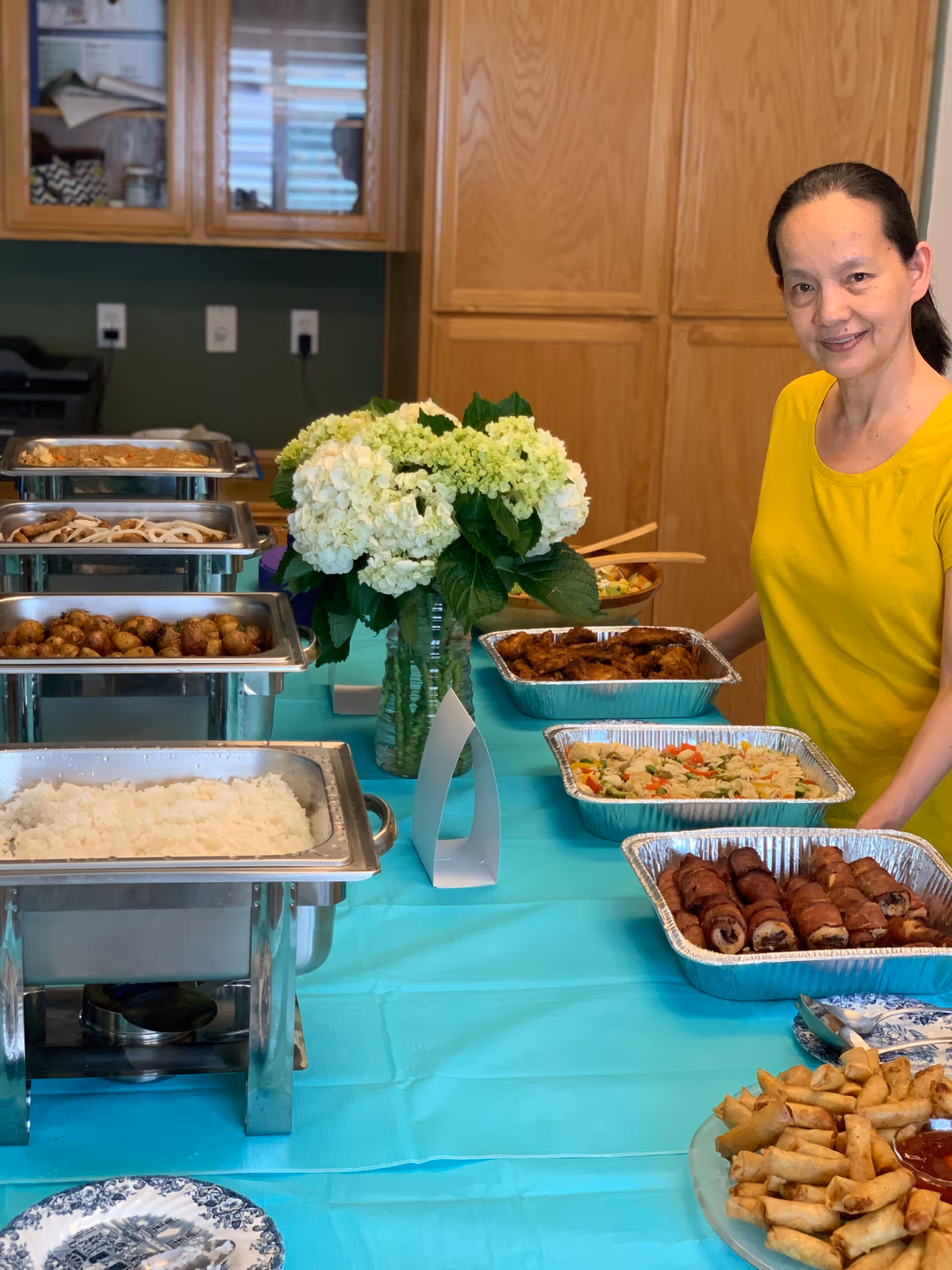 A woman in a yellow shirt stands behind a buffet table covered with a turquoise tablecloth. The table holds several trays of food including rice, roasted potatoes, spring rolls, and other dishes. A vase with white and green hydrangea flowers is placed in the center of the table. Wooden cabinets and kitchen appliances are visible in the background.