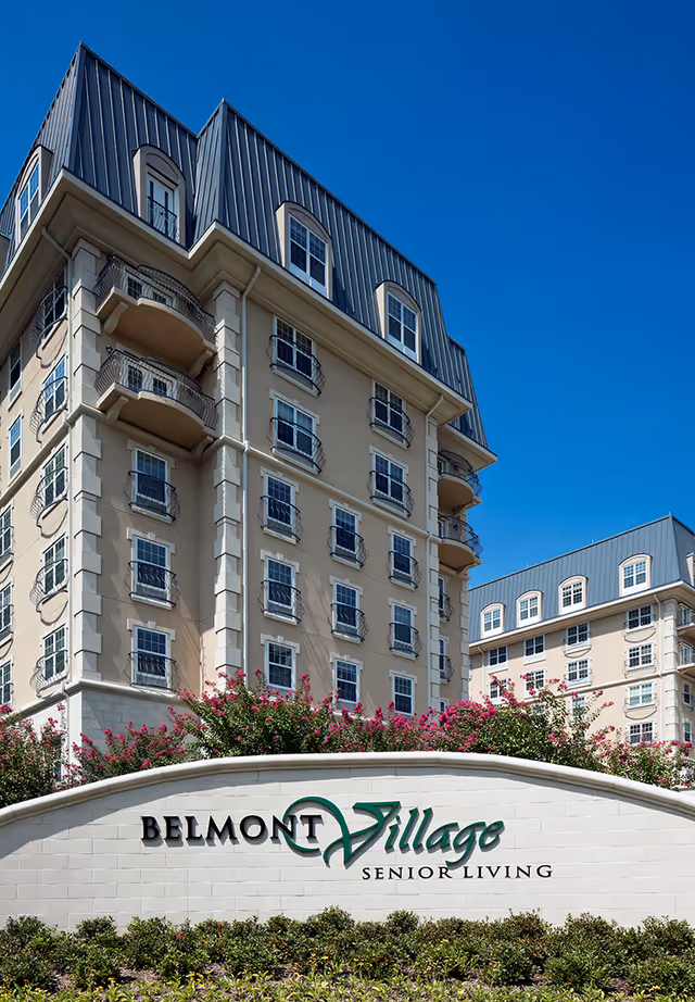 Exterior view of Belmont Village Senior Living Turtle Creek, showing a multi-story beige building with multiple windows and balconies under a clear blue sky. In front, there is a white stone sign with the facility's name and landscaping with green bushes and pink flowers.