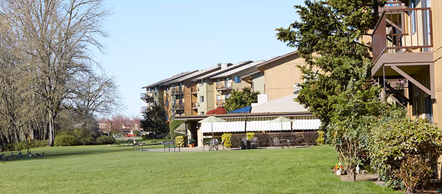 A large green lawn with several benches and a few geese scattered around. In the background, there is a multi-story residential building with balconies and a covered patio area. Trees line the left side of the lawn under a clear blue sky.