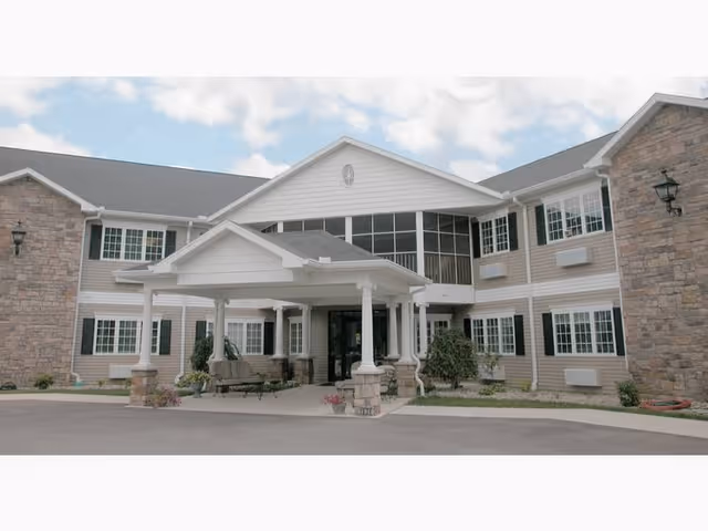 Front entrance of a two-story senior living building with a covered porte-cochere, stone accents, and multiple windows.