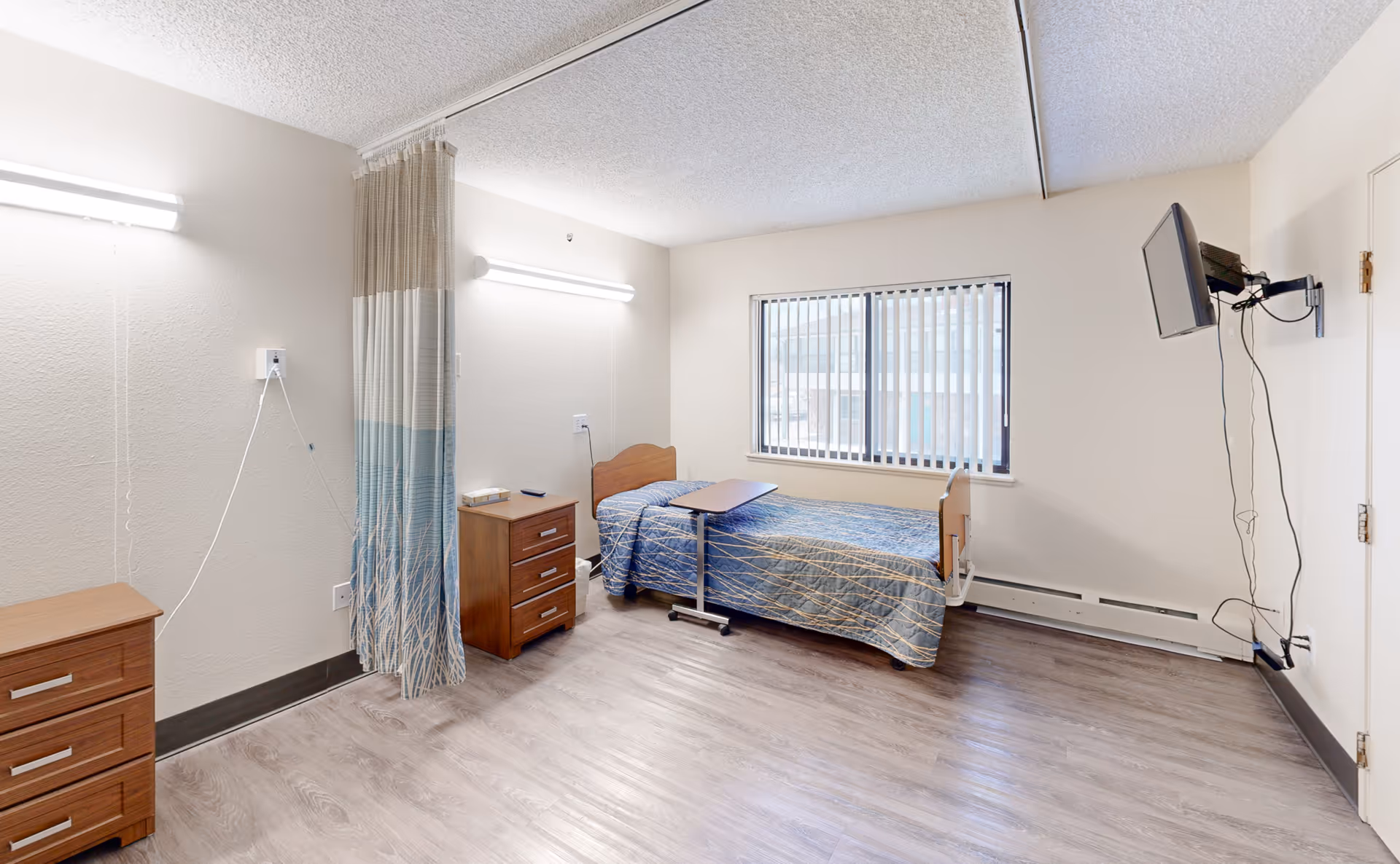 A simple, clean room in a rehabilitation and care center featuring a single bed with a blue patterned bedspread, a small overbed table, two wooden nightstands, a wall-mounted television with visible cords, a window with vertical blinds, and a privacy curtain partially drawn along one wall. The room has light-colored walls and wood-look flooring.