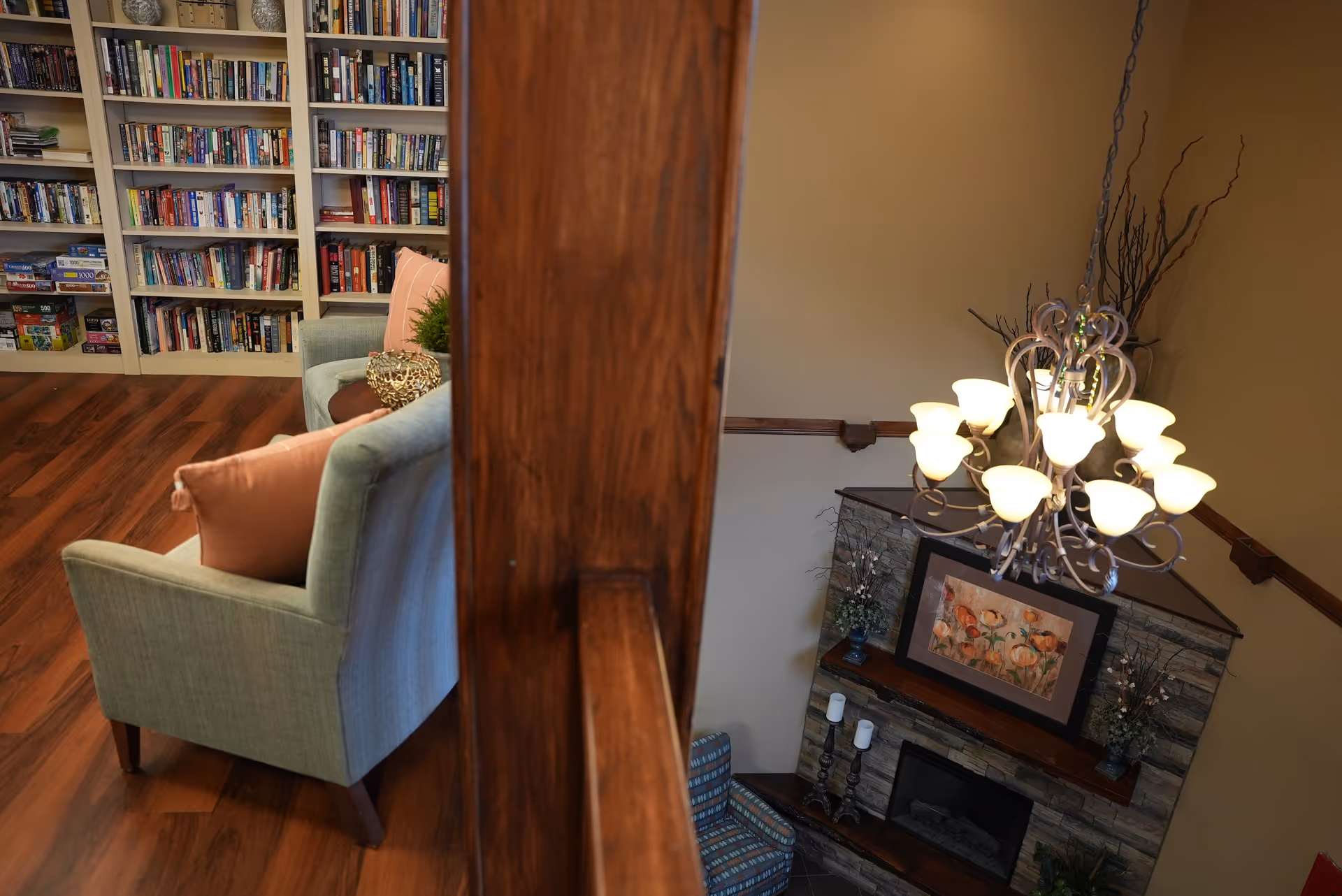 Interior view of a senior living facility showing a cozy reading area with bookshelves filled with books and board games, a light blue armchair with a pink pillow, and a decorative centerpiece on a small table. The image also captures a view down to a living room area with a stone fireplace, framed artwork, candles, and a chandelier with multiple lights hanging from the ceiling.