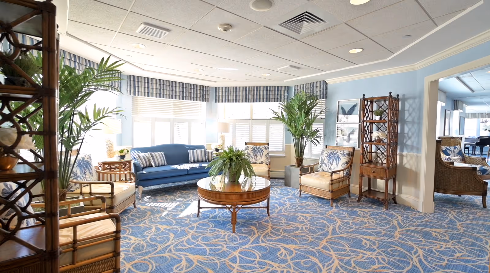 A bright and airy living room with blue walls and patterned blue carpet. The room features a blue sofa with striped cushions, two armchairs with palm tree patterned cushions, a round wooden coffee table with a green plant centerpiece, and tall green plants near the windows. The windows have white blinds and blue plaid valances. There is a wooden shelving unit on the right side and an open doorway leading to another room with similar decor.