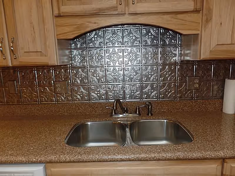 Double stainless steel kitchen sink with a faucet set into a brown countertop, wood cabinets above and a decorative metallic tile backsplash.