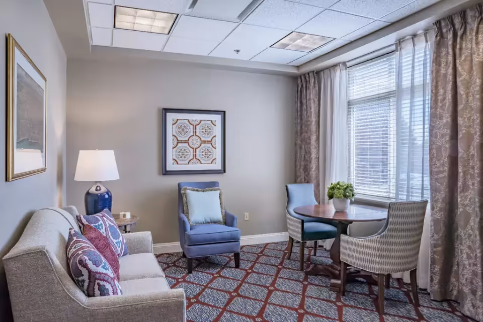 Cozy sitting room with a sofa, patterned pillows, an armchair, a table lamp, and a round table with chairs by a large window.