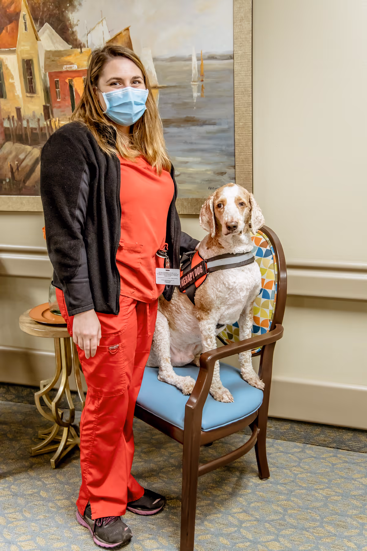 A woman wearing a red uniform and a face mask stands next to a therapy dog sitting on a blue cushioned chair in an indoor setting. Behind them is a painting of a waterfront scene with boats and buildings.