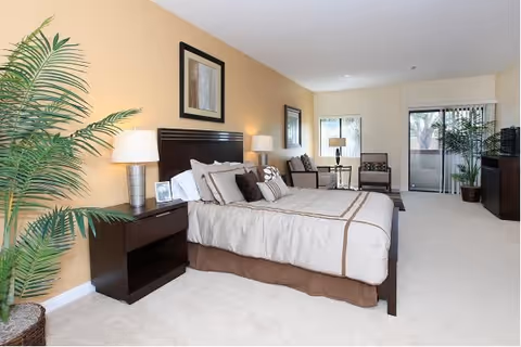 A spacious bedroom with a large bed featuring beige and brown bedding, two bedside tables with lamps, a framed picture above the bed, a sitting area with chairs and a small table near a window, a sliding glass door leading to a balcony, and a large potted plant in the corner.