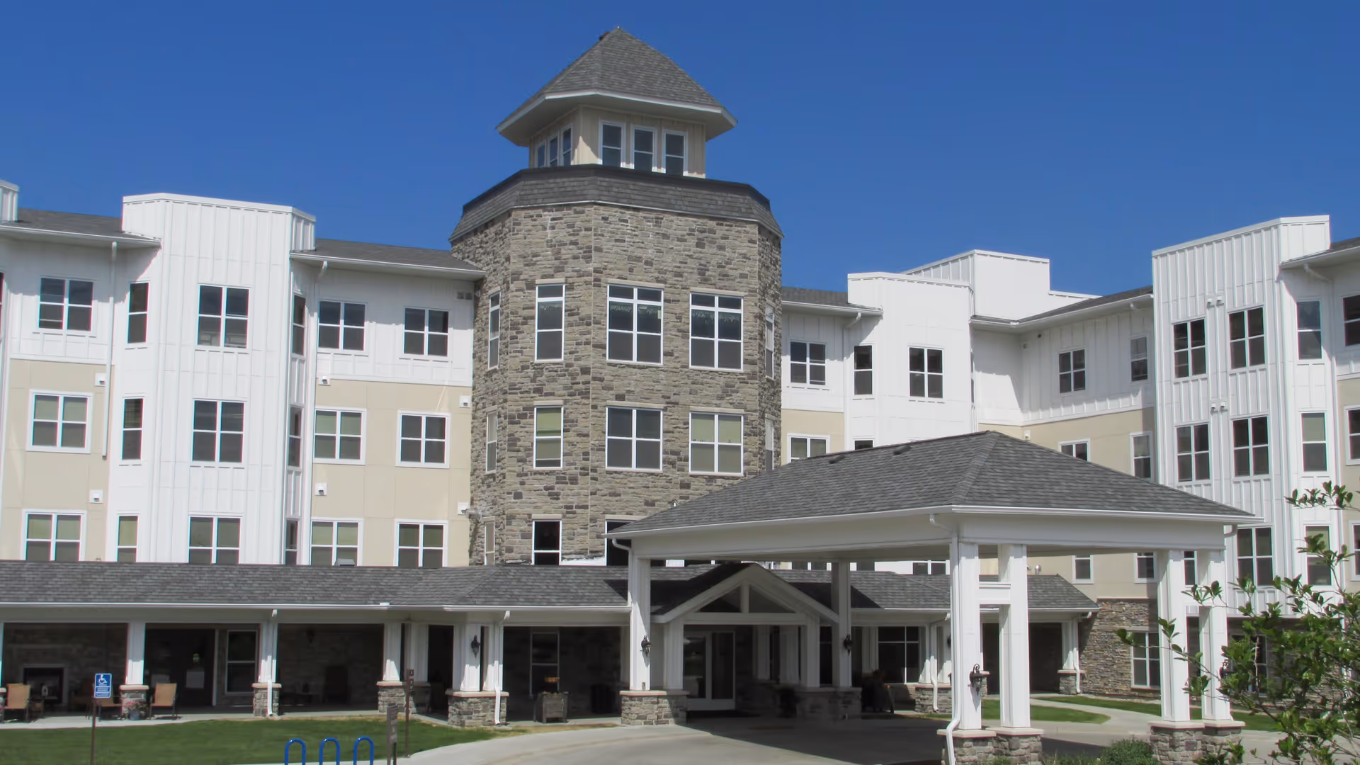 Front exterior view of York Gardens facility showing a multi-story building with a stone tower in the center, white siding, numerous windows, and a covered entrance driveway under a clear blue sky.