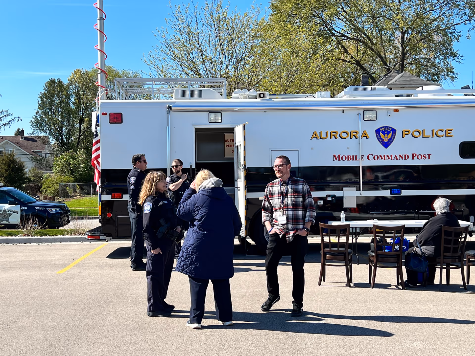 Several people, including police officers and civilians, standing and sitting near an Aurora Police Mobile Command Post vehicle parked in an outdoor area on a sunny day with trees and houses in the background.