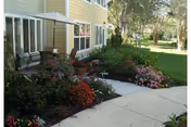 Sidewalk leading past a landscaped flower garden and a small patio area beside a yellow residential building.