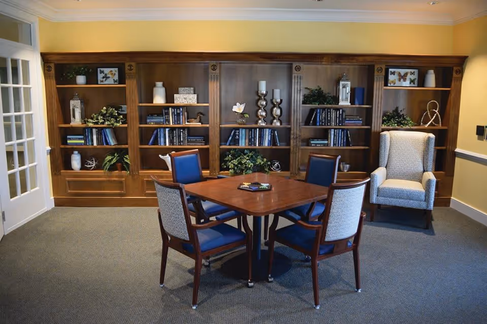A cozy room with a wooden table surrounded by four chairs, two with blue cushions and two with patterned cushions. Behind the table is a large wooden bookshelf filled with books, decorative plants, candles, and framed butterfly artwork. To the right of the bookshelf is a comfortable upholstered armchair. The walls are painted yellow and the floor is carpeted.