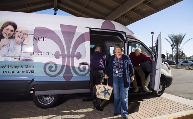A woman carrying a tote bag is walking away from a white shuttle van with the Renaissance Village Murrieta logo on its side. Two other people are seen exiting the van in a parking lot area with palm trees in the background.