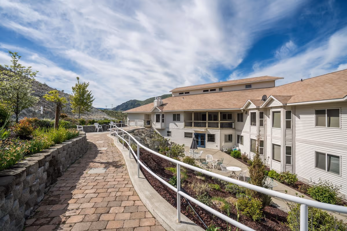 Paved outdoor walkway and patio with tables and chairs next to landscaped beds leading to a multi-story white senior living building under a partly cloudy sky.