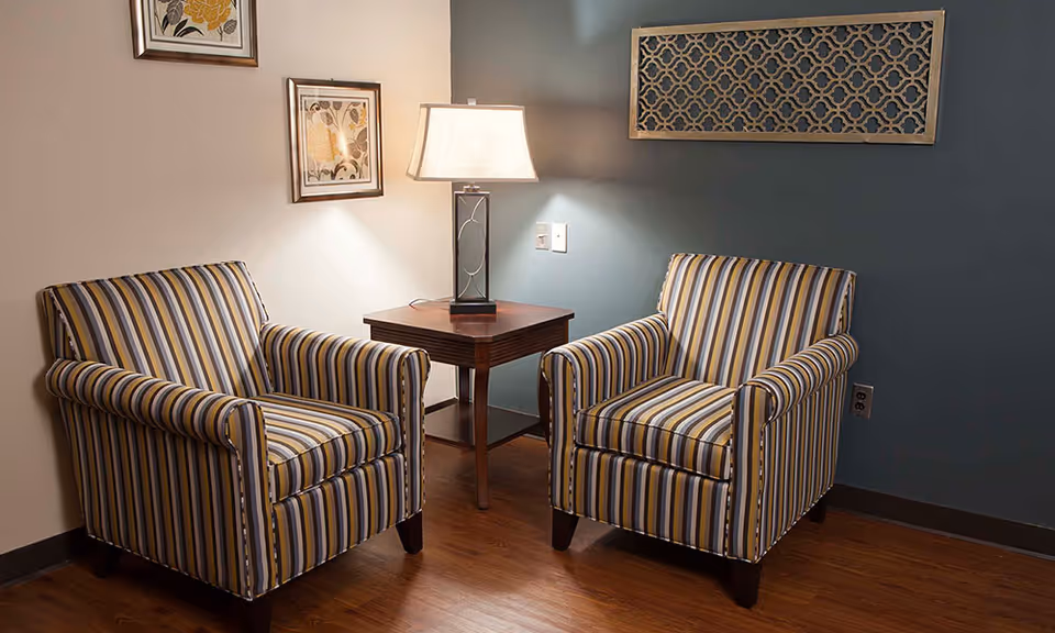Two striped armchairs with yellow, gray, and white colors are placed on either side of a wooden side table with a lamp on it. The room has wooden flooring, a blue accent wall with a decorative wall piece, and two framed floral artworks on the adjacent wall.