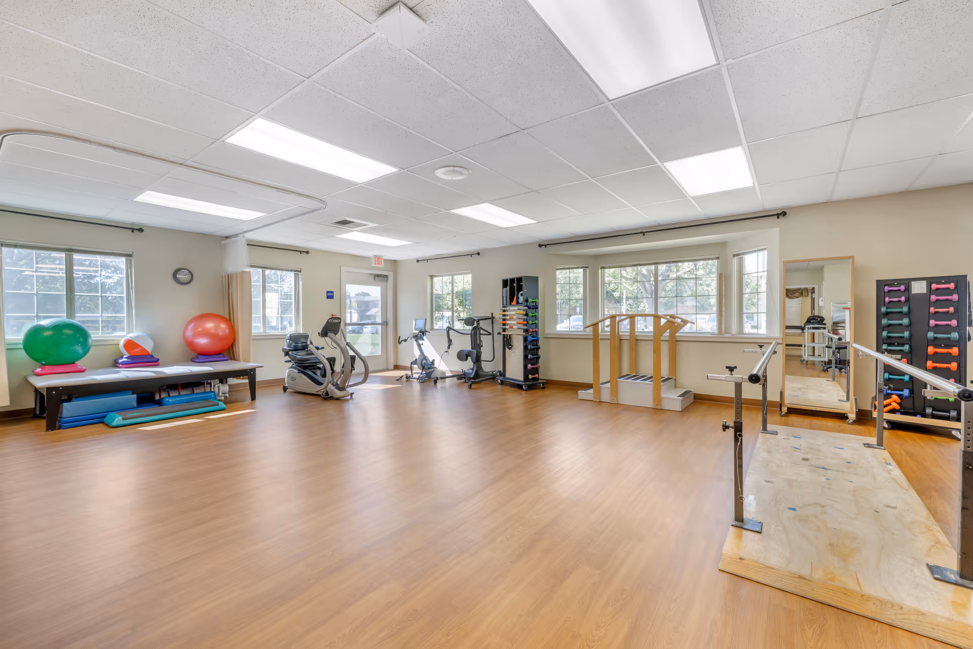A spacious physical therapy or exercise room with wooden flooring, large windows letting in natural light, exercise equipment including stationary bikes, balance balls, hand weights, parallel bars, and a wooden step platform. The room has a white ceiling with fluorescent lights and beige walls.