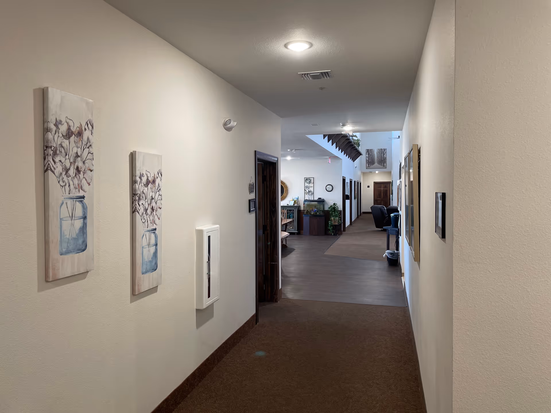 A hallway inside a senior living facility with beige walls and brown carpet. The hallway leads to a common area with seating and decorative plants. There are framed artworks on the walls and ceiling lights illuminating the space.