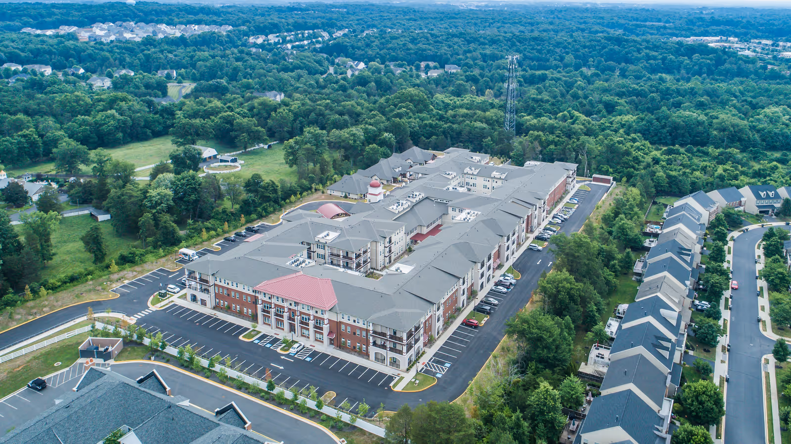 Aerial view of a large multi-building senior living complex with parking lots, surrounded by trees and nearby houses.