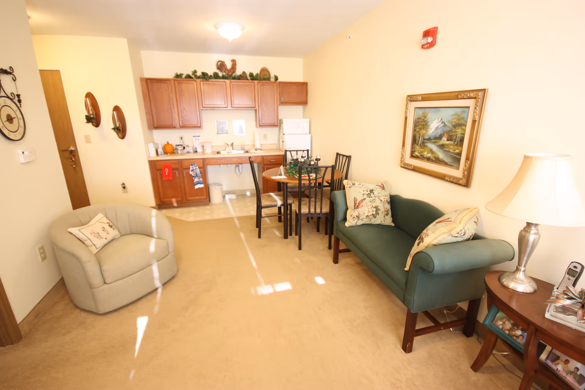 A cozy living area in a senior living facility featuring a green upholstered loveseat with floral pillows, a round wooden dining table with four chairs, a beige armchair with a floral pillow, and a small wooden side table with a lamp and a cordless phone. The background shows a kitchenette with wooden cabinets, a microwave, a sink, and some decorative items including a rooster figurine and greenery on top of the cabinets. The walls are light-colored and there is a framed painting of a mountain landscape hanging above the loveseat.