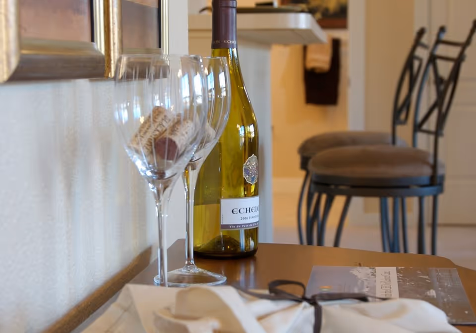 Close-up of a table with two empty wine glasses containing wine corks, a bottle of Echelon 2006 Pinot Noir wine, and a folded napkin with a ribbon. In the background, there are cushioned bar stools and part of a kitchen counter.