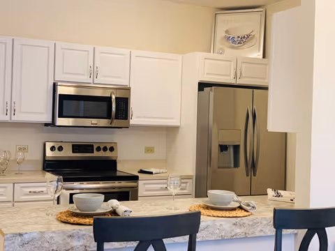 Modern kitchen with white cabinets, stainless steel refrigerator, oven, and microwave. A granite countertop with place settings including bowls, glasses, and napkins is visible in the foreground with black chairs.